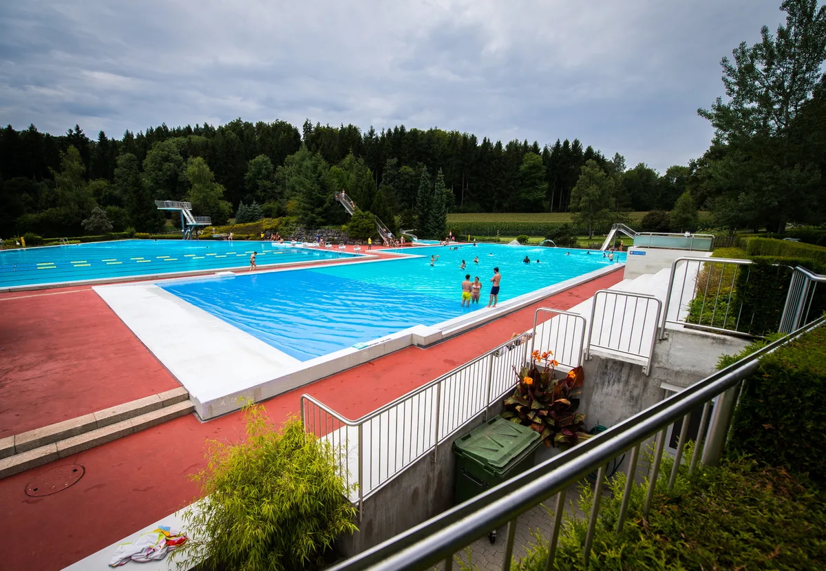 Blick auf ein Freibad mit zwei Becken, in denen sich mehrere Personen befinden. 